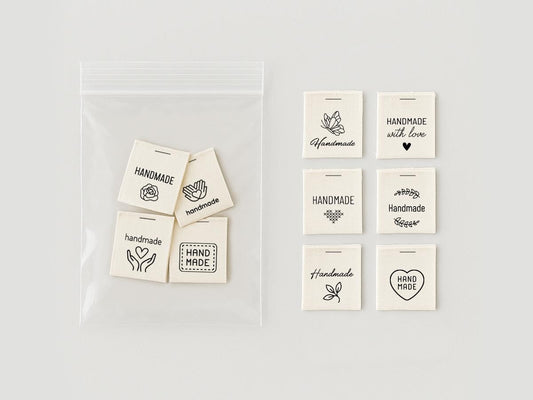 May include: A clear plastic bag containing several off-white fabric tags with the word "HANDMADE" and various small black illustrations. Additional tags with "HANDMADE" and "HAND MADE" are displayed separately.